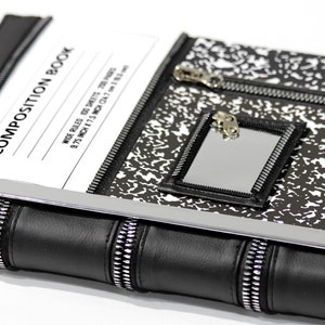 May include: A black and white composition book cover featuring a speckled pattern, a rectangular mirror with a zipper border, and a white label with the text COMPOSITION BOOK, WIDE RULED, 100 SHEETS, 200 PAGES, 9.75 INCH X 7.5 INCH (24.7 cm X 19.1 cm).
