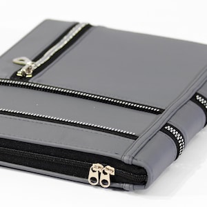 May include: A rectangular gray portfolio case featuring multiple horizontal black and silver zippers across the front surface. The case includes a main black zippered closure along the side with two silver metal pull tabs for access.