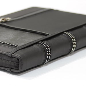 Könnte beinhalten: Schwarzes Leder-Portfolio mit silbernen Reißverschlüssen. Das Portfolio hat einen Klappenverschluss und einen Reißverschluss, der um den gesamten Umfang verläuft. Das Design umfasst eine strukturierte Lederplatte und ein silbernes Reißverschlussdetail am Rücken.