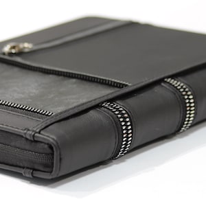 May include: A black portfolio case featuring a smooth matte finish, a zippered main compartment, and a front pocket with a silver-toned metal zipper. The side edge displays two decorative vertical zipper accents with metallic teeth.