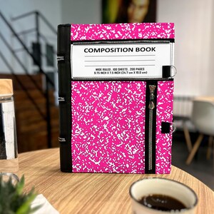 May include: A bright pink and white speckled composition book cover featuring a black spine, a horizontal zipper across the top, and a vertical zipper on the right side. The label reads COMPOSITION BOOK, WIDE RULED, 100 SHEETS, 200 PAGES, 9.75 INCH X 7.5 INCH.