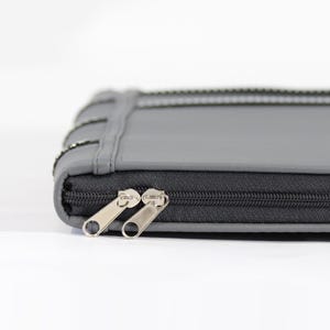 May include: A rectangular gray case with a smooth matte finish and a black zipper closure. The case features two silver-toned metal zipper pulls and a stitched seam detail running along the side edge of the exterior surface.