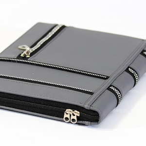 May include: A gray portfolio with multiple black zippers. The portfolio has a rectangular shape and a silver zipper pull. The zippers are placed on the front and sides of the portfolio. The background is white.