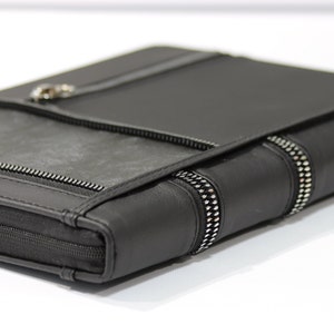 Op de afbeelding: Zwart leren portfolio met een zilveren ritssluiting en een flap. De portfolio heeft een getextureerd, donkergrijs gedeelte aan de voorkant en een zilveren metalen sluiting. De randen zijn geaccentueerd met zilveren studs.