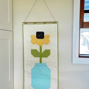 May include: A rectangular fabric wall hanging featuring a quilted design of a yellow flower with a black centre in a light blue mason jar. The piece is set against a cream background with a green border, suspended from a wooden dowel with a white cord.