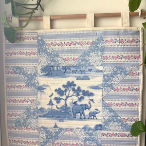 May include: A square fabric wall hanging featuring a central panel with a blue and white toile print of elephants and trees. The border consists of pieced fabric blocks in blue and white stripes with small pink floral accents, suspended from a wooden rod.