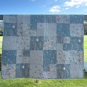 May include: A patchwork quilt featuring a variety of blue fabric squares. The design includes alternating blocks of solid light blue with swirling quilted patterns, blue and white gingham checks, and a print depicting small illustrated rabbits.