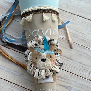 Könnte beinhalten: Eine beige und blaue Schultüte mit dem Namen "David" auf der Vorderseite. Die Tüte ist mit einem Löwenkopf mit türkisfarbenem Stirnband und Feder verziert. Das Datum "16.08.2025" ist unten eingestickt.