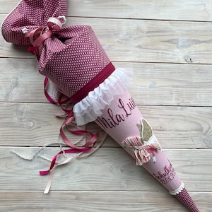 Peut inclure: Un cône d'école rose et blanc avec une bordure en dentelle blanche et un ruban rose. Le cône est décoré d'une licorne pailletée dorée et le nom "Mila Lu" est écrit en rose. Le cône porte également le texte "Schulkind 2023" écrit en rose.