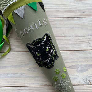 May include: A green and white striped school cone featuring a black panther patch and the name "leonis" embroidered in white. The cone has green paw prints and the text "Schulkind 2025". The top is decorated with small felt flags.