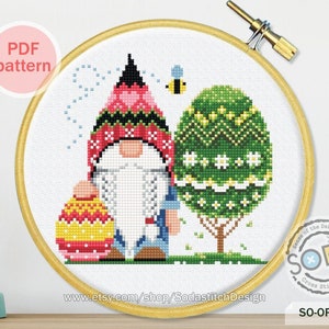 Könnte beinhalten: Kreuzstichmuster mit einem Zwerg, der ein buntes Osterei hält, neben einem grünen Baum mit weißen Blüten und einer gelben Biene. Das Muster trägt den Titel "SO-OP188" und ist als PDF-Download erhältlich.