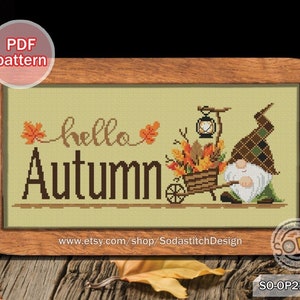 May include: Cross stitch pattern featuring a gnome with a wheelbarrow full of autumn leaves and a lantern. The text "hello autumn" is stitched above the gnome. The pattern is framed in a brown wood frame.