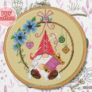 Puede incluir: Patrón de punto de cruz con un gnomo sentado con un carrete de hilo y una aguja, rodeado de flores, botones y un corazón. El patrón se titula "SO-OP259" y está disponible para descargar en PDF.