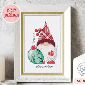 May include: Cross stitch pattern featuring a gnome holding a turquoise ornament with the word "December" and the text "Turquoise" written vertically. The gnome is wearing a red and white hat with a white beard and red boots. The pattern is labeled "SO-BS12".