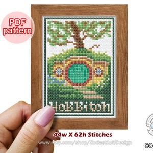 May include: A cross-stitch pattern of a green hobbit house with a round green door, surrounded by green grass and a tree. The pattern is framed in wood and has the text "Hobbiton" below the house. The pattern is 44 stitches wide and 62 stitches high.