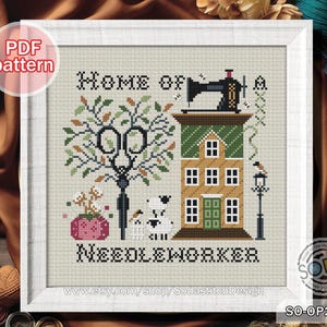 Könnte beinhalten: Gerahmtes Kreuzstichmuster in Weiß. Das Design zeigt ein Haus mit einer Nähmaschine, einen Baum mit einer Schere und den Text "HOME OF NEEDLEWORKER". Ein Hund und ein Vogel sind ebenfalls im Design enthalten.