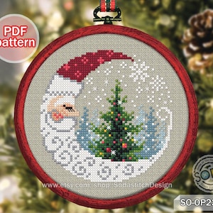 May include: A cross-stitch Christmas ornament featuring Santa Claus in a crescent moon shape, with a Christmas tree and snowflakes. The ornament is framed in a red hoop and has a PDF pattern label.