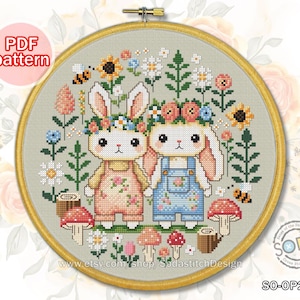 Può includere: Schema di punto croce con due coniglietti adorabili che indossano corone di fiori e salopette, circondati da fiori e funghi colorati. Lo schema è in un cerchio e include il testo "SodastitchDesign".