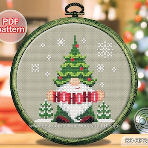 May include: Cross stitch pattern featuring a gnome wearing a green Christmas tree hat and a red scarf with the word "HO HO HO" on it. The gnome is standing in front of a small green tree and snowflakes.