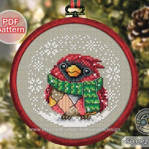 May include: A cross-stitch Christmas decoration featuring a red bird wearing a green scarf, surrounded by snowflakes. The ornament is set in a red hoop with a green and red ribbon for hanging. The text "PDF pattern" is visible.