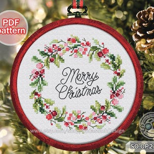 May include: A circular cross-stitch embroidery in a red frame, featuring a Christmas wreath. The wreath is composed of green leaves and red berries, encircling the words "Merry Christmas" in black script. A PDF pattern is indicated.