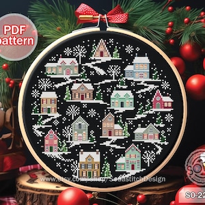 May include: A black and white cross stitch pattern of a winter village scene with houses, trees, and snowflakes. The pattern is framed in a wooden hoop and surrounded by Christmas decorations.