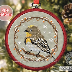May include: A cross-stitch pattern for a Christmas ornament featuring a small bird with yellow and brown feathers perched on a branch with berries and snowflakes. The pattern is in a red hoop with the text "SO-OP2262" and "www.etsy.com/shop/SodastitchDesign" at the bottom.