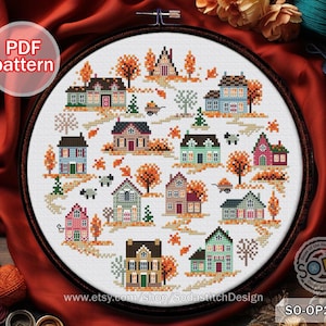 May include: Cross stitch pattern depicting a charming autumnal village scene with houses, trees, and a whimsical cart. The pattern is in a hoop and includes the text "SO-OP2242" and "www.etsy.com/shop/SodastitchDesign".