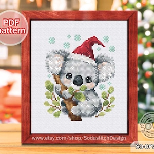 May include: Cross-stitch pattern of a koala wearing a Santa hat, holding a branch with green leaves, and surrounded by snowflakes. The artwork is framed in a red wooden frame. Text on the frame reads "PDF pattern" and "www.etsy.com/shop/SodastitchDesign".