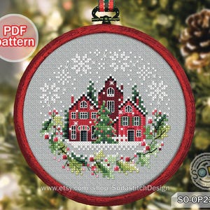 Könnte beinhalten: Ein Kreuzstich-Weihnachtsschmuck mit einer rotbedachten Dorflandschaft mit einem Weihnachtsbaum, Schneeflocken und einem grünen Kranz. Das Design befindet sich in einem runden, roten Rahmen mit einem Aufhängeband.