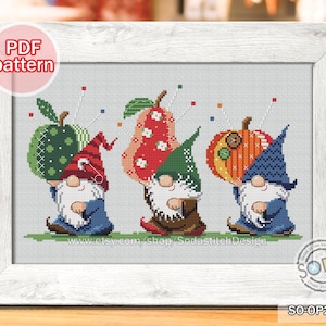 May include: Cross-stitch pattern featuring three gnomes holding a green apple, a red pear, and an orange pumpkin. Each gnome wears a different colored hat and holds a fruit with a needle and thread. The image is framed in a white wooden frame.