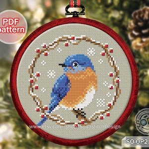 May include: A bluebird cross stitch pattern in a red embroidery hoop with a white background. The bird is surrounded by a wreath of brown branches with red berries and snowflakes. The pattern is titled "SO-OP2136".