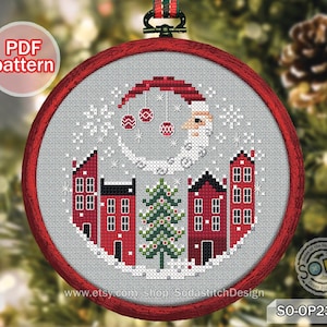 May include: A cross-stitch Christmas ornament featuring a winter village scene with red houses, a Christmas tree, and Santa Claus. The design is set within a red circular frame. The text "PDF pattern" is visible.