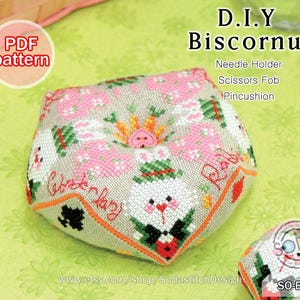 May include: A PDF pattern for a DIY biscornu, a small, round, stuffed cushion. The pattern features a whimsical design with bunnies, flowers, and hearts. The biscornu can be used as a needle holder, scissors fob, or pincushion. The pattern is available for download on Etsy.