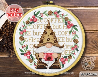 May include: A cross-stitch pattern in a wooden hoop features a gnome holding a coffee cup. The gnome wears a brown hat with coffee bean accents. The text reads "But first COFFEE" surrounded by a wreath of coffee beans and berries. The PDF pattern is available on Etsy.