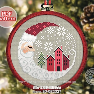 Può includere: Uno schema a punto croce in un telaio rotondo in legno rosso che raffigura una mezzaluna a forma di Babbo Natale addormentato, un piccolo pino verde e tre case rosse. Il disegno include fiocchi di neve bianchi su fondo in tessuto beige.