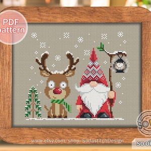 以下が含まれることがあります： 赤い帽子と白いひげのノームが提灯を持ち、緑色のマフラーをしたトナカイが隣にいるクロスステッチのパターンです。ベージュの背景に雪の結晶が散りばめられています。