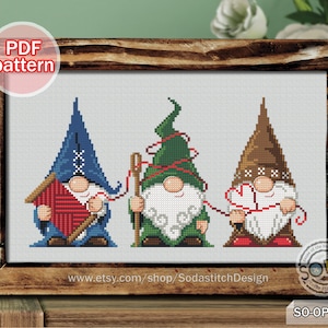 Puede incluir: Patrón de punto de cruz con tres gnomos con sombreros rojo, verde y azul. Los gnomos sostienen hilo y agujas, lo que sugiere un tema artesanal. El patrón se titula "SO-OP2213" e incluye el sitio web "www.etsy.com/shop/SodastitchDesign".
