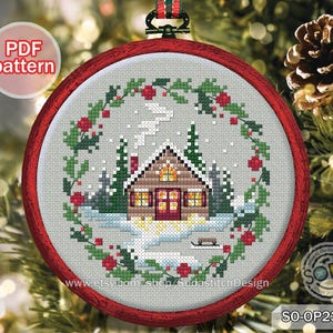May include: A cross-stitch embroidery in a round red frame, featuring a winter cabin scene. The cabin has lit windows, snow on the roof, and a wreath of greenery and red berries. The text "PDF pattern" is in the upper left.