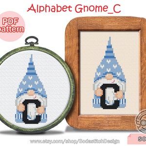 Può includere: Schema di punto croce di un gnomo che tiene la lettera C. Il gnomo indossa un cappello blu con cuori bianchi e una barba bianca. La lettera C è nera. Lo schema è in una cornice di legno con sfondo bianco.