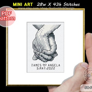 May include: Black and white cross stitch pattern of two hands holding each other. The text below reads "JAMES ANGELA 3.MAY.2022".