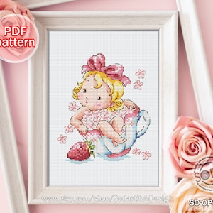 Op de afbeelding: Borduurpatroon met een schattig cartoon babymeisje dat in een theekopje zit met een roze strik en een aardbei. Het patroon is op een witte achtergrond met roze bloemen.