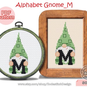 Puede incluir: Patrón de punto de cruz de un gnomo verde sosteniendo la letra M. El patrón está en un marco de madera y está etiquetado como "Alphabet Gnome_M" y "SO-M".