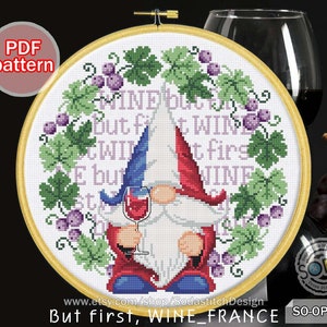 May include: Cross stitch pattern featuring a gnome with a red hat and blue pants holding a glass of wine. The gnome is surrounded by a wreath of green leaves and purple grapes. The text "MIND BUL BUT f WIL E but WIN at firs WINE" is stitched around the gnome. The text "But first, WINE_FRANCE SO-OP2160" is at the bottom of the pattern.