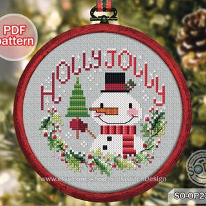 Può includere: Un motivo a punto croce in un cerchio rosso presenta un pupazzo di neve con un cappello a cilindro nero e una sciarpa rossa, circondato dalle parole "HOLLY JOLLY" e da vegetazione festiva. Il design include un albero di Natale verde e un uccello rosso. Il testo "PDF pattern" è visibile.