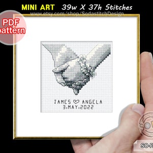 May include: A black and white cross-stitch pattern of two hands holding each other. The pattern includes the text "JAMES ANGELA 3.MAY.2022". The pattern is 39 stitches wide and 37 stitches high.