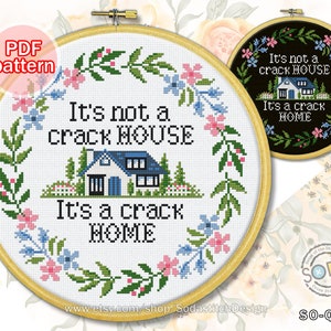 May include: Cross stitch pattern for a hoop with a white background and a floral wreath surrounding the text "It's not a crack house, It's a crack home" with a small house illustration in the centre.