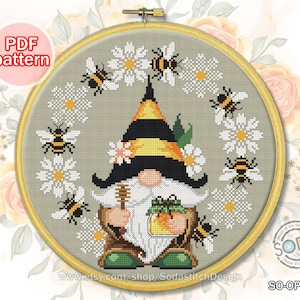 May include: A cross-stitch pattern featuring a gnome holding a jar of honey surrounded by bees and daisies. The pattern is in a round hoop and is titled "SO-OP2162".