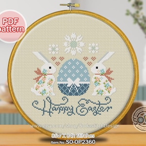 Puede incluir: Un bordado de punto de cruz en un aro de madera presenta un huevo de Pascua azul entre dos conejitos blancos con motivos florales. El texto "Happy Easter" está bordado debajo. El diseño incluye el texto "PDF pattern".