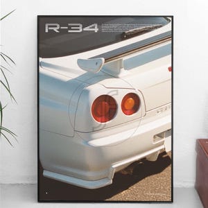 Könnte beinhalten: Poster mit einem weißen Auto und dem Text "R-34". Das Bild zeigt das Heck des Fahrzeugs, einschließlich der Rücklichter und des Spoilers. Das Poster ist in Schwarz gerahmt und vor einem neutralen Hintergrund platziert.